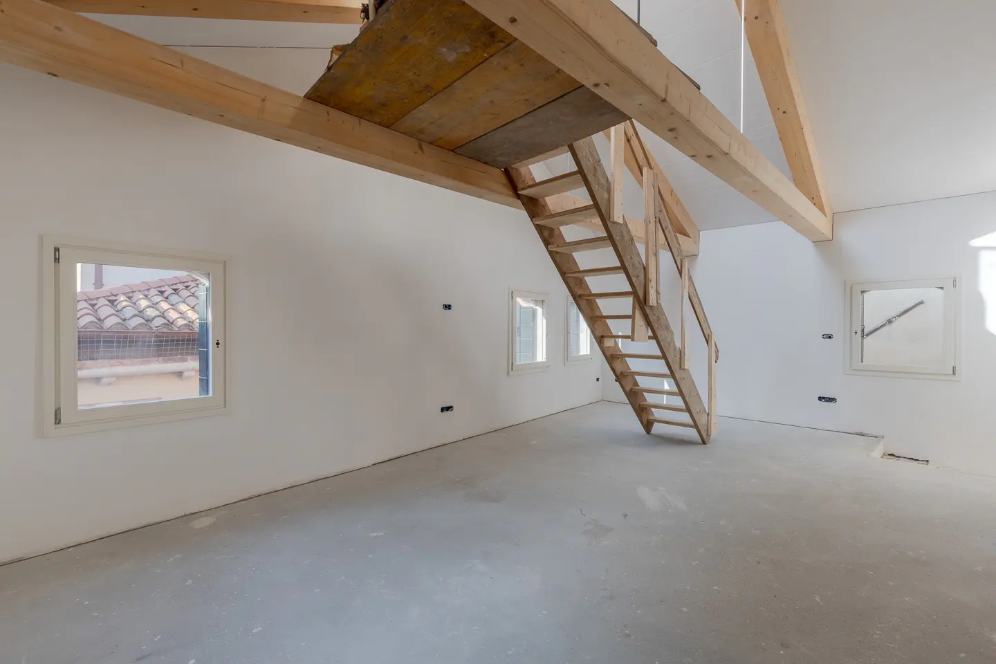 Unfinished room with white walls, gray floor, and wooden stairs leading to a loft. Windows show rooftops outside.