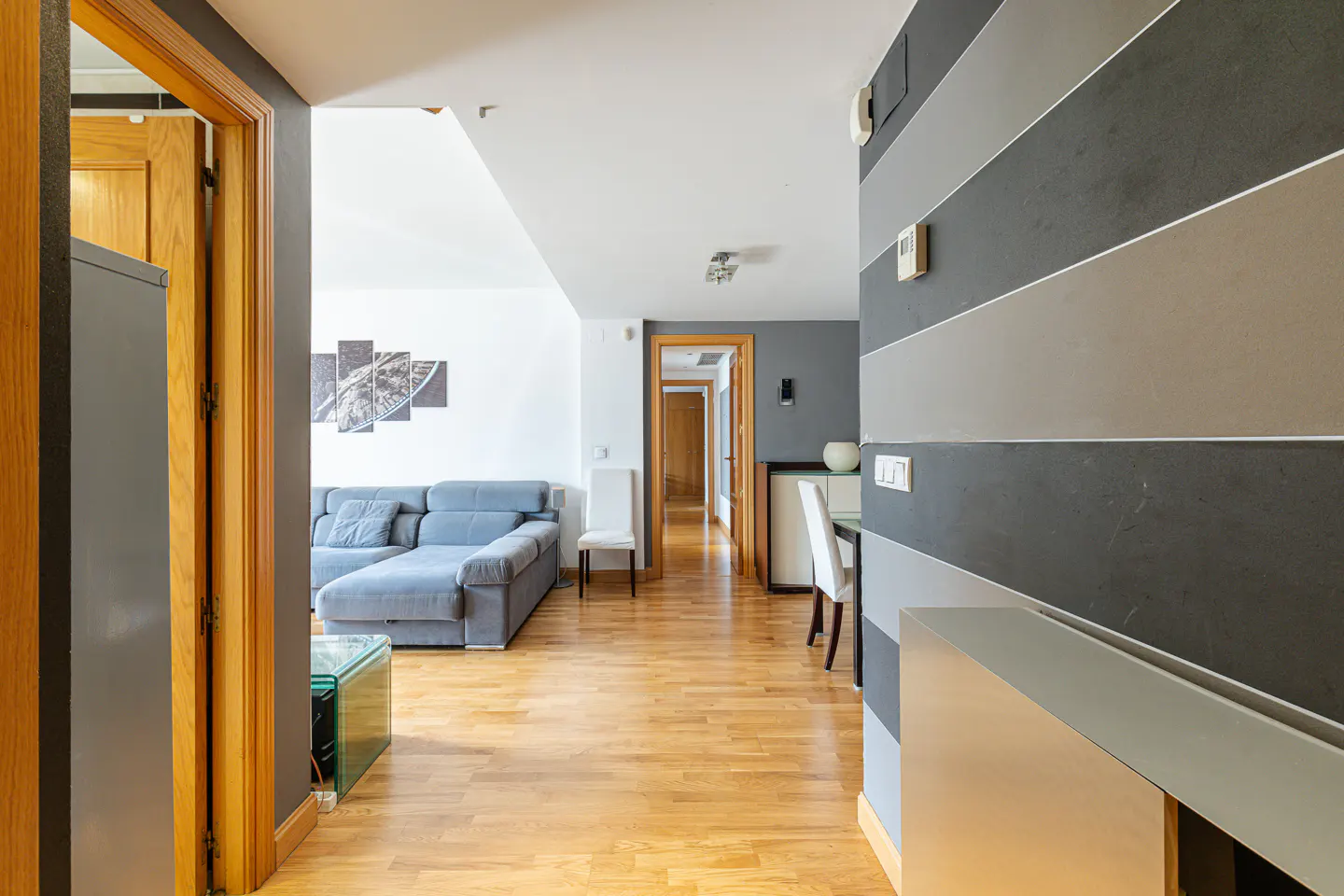 A modern apartment hallway with wood floors, a gray sofa, and gray and white striped walls.