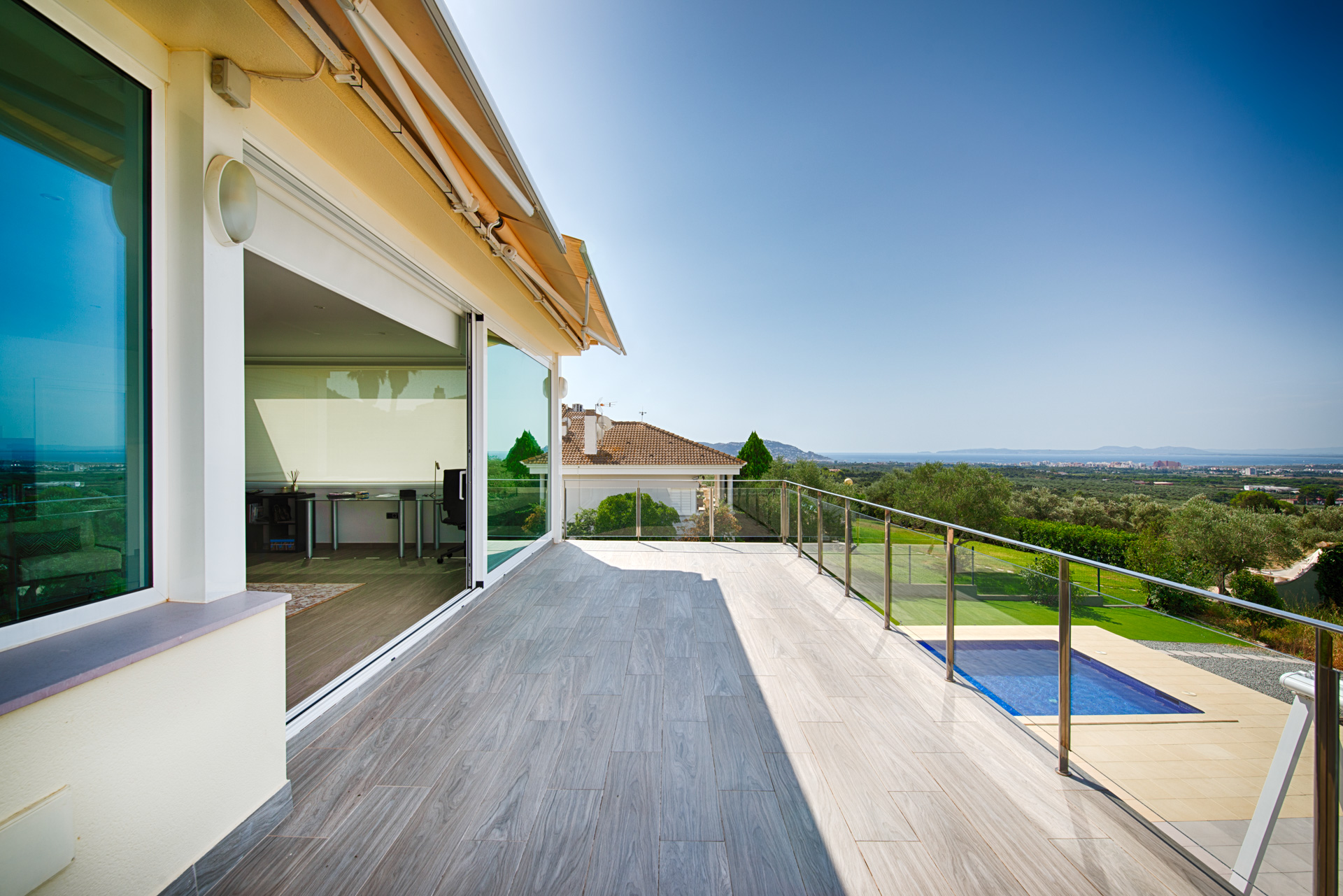 Spacious balcony with wood-look tile, glass railing, and a view of a swimming pool, garden, and distant coastline. [AI-generated text]