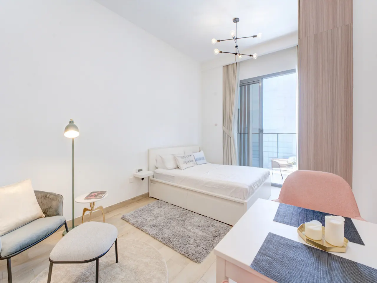 Bright studio apartment with white walls, a bed, a chair, and a dining table near a balcony.
