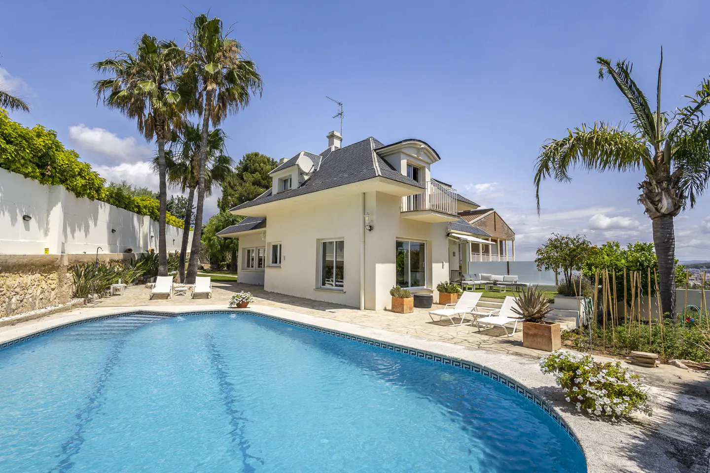 A cream-colored two-story house with a dark roof, featuring a blue swimming pool, palm trees, and lounge chairs on a patio.