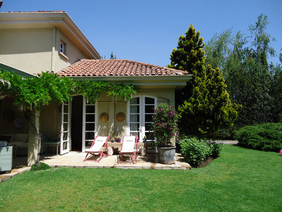 A tan house with a red tile roof has a patio with two white lounge chairs. Green grass and trees surround the house.