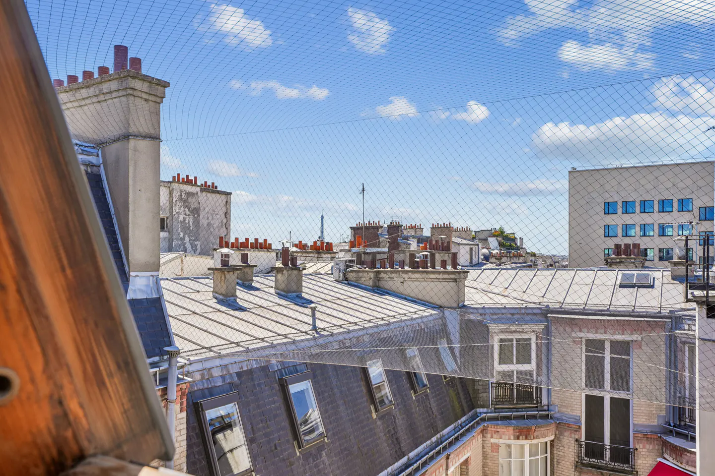 View of Paris rooftops with chimneys and dormer windows, seen through a safety net against a blue sky.