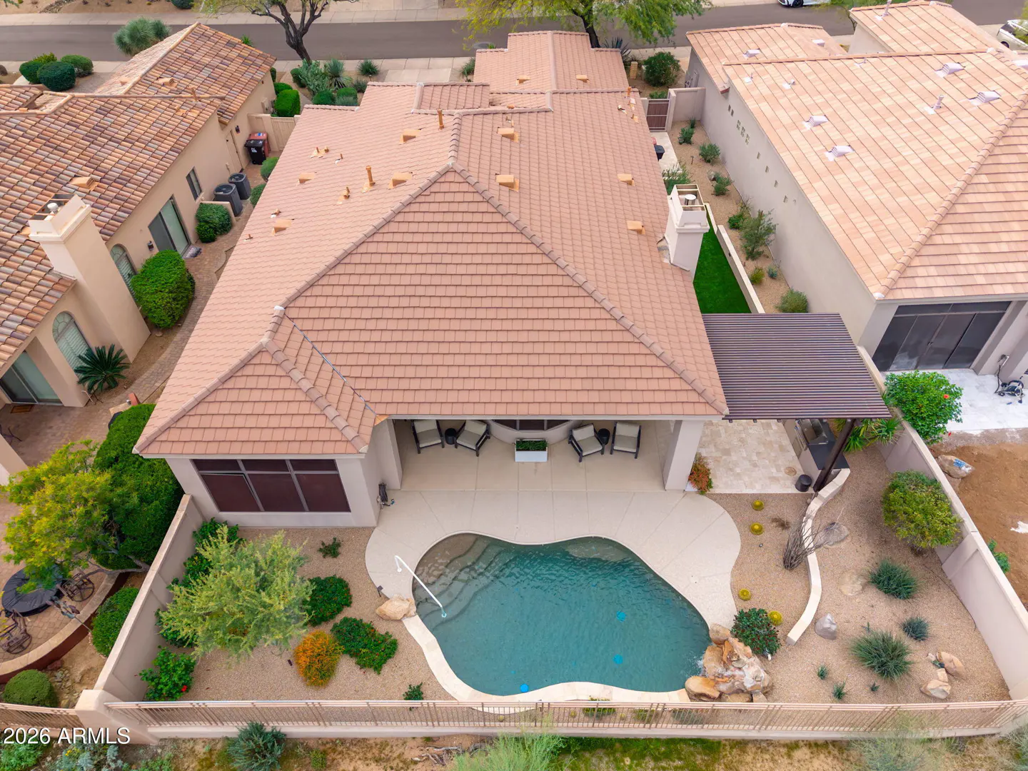Aerial view of a tan house with a brown tile roof, a pool, and a covered patio with chairs.