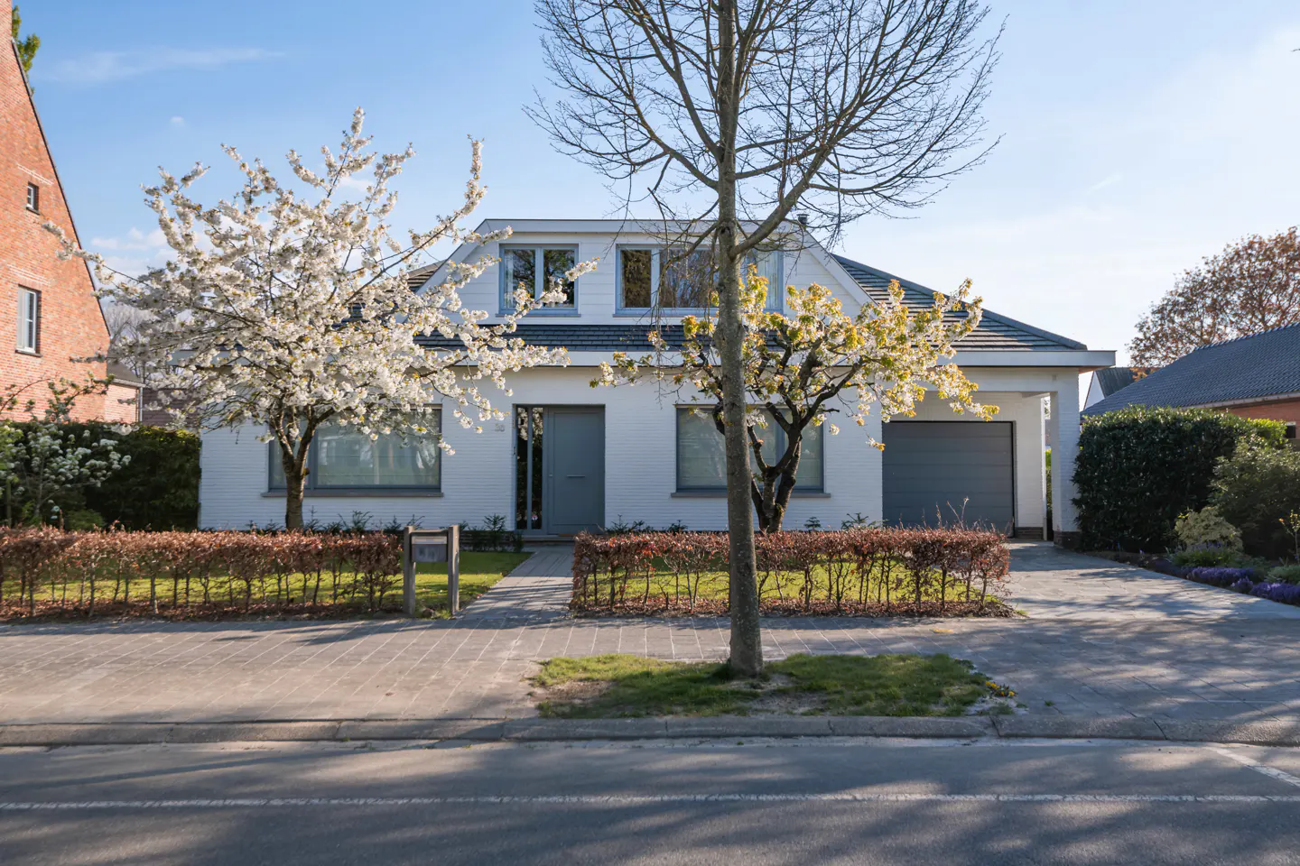 A white house with a gray door and garage is framed by flowering trees and a trimmed hedge.