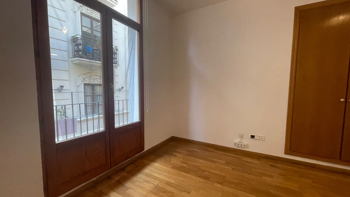 A bright, empty room with hardwood floors, a brown wardrobe, and a window with a view of a building with balconies.