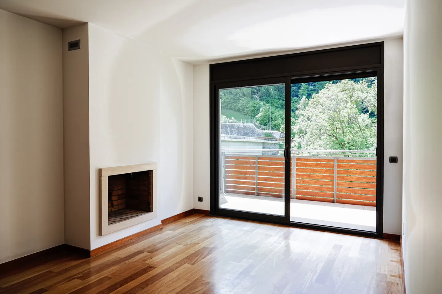Bright, empty room with hardwood floors, white walls, and a stone fireplace. Sliding glass doors open to a balcony with a wood railing and a view of trees.