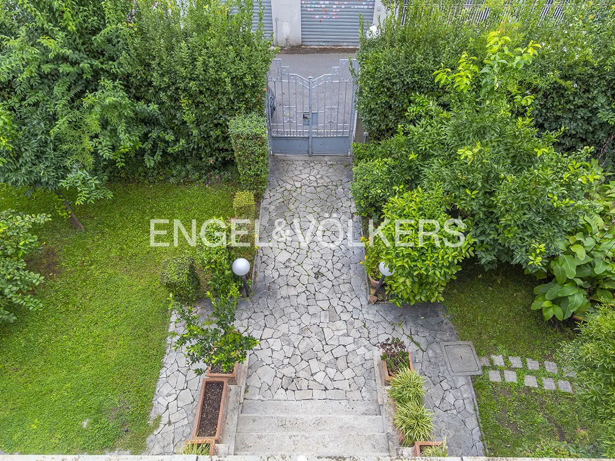 Aerial view of a stone path leading to a gray gate, surrounded by green bushes and grass. The Engel & Völkers logo is superimposed on the image.