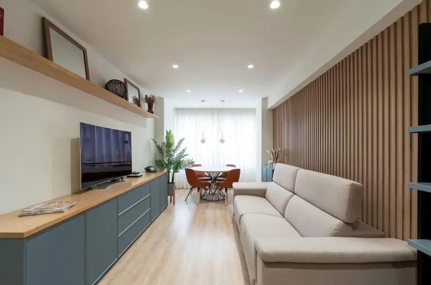 Bright living room with a beige sofa, a round table with orange chairs, and a large TV on a blue cabinet.