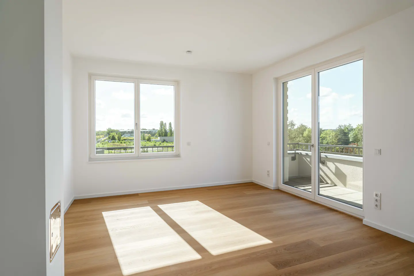 Empty room with white walls, light wood floors, a window, and a glass door leading to a balcony with a green view.