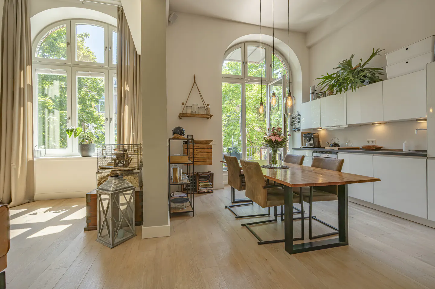 Bright, modern apartment interior with wood floors, arched windows, and a dining table with four chairs. White kitchen cabinets line the back wall.