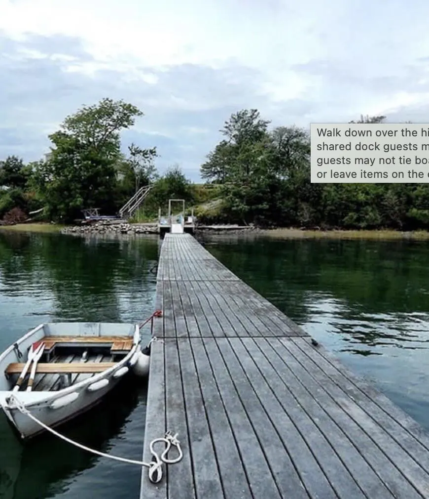 A long wooden dock extends into calm, green water, with a small white rowboat tied to its side. Trees line the shore under a cloudy sky.