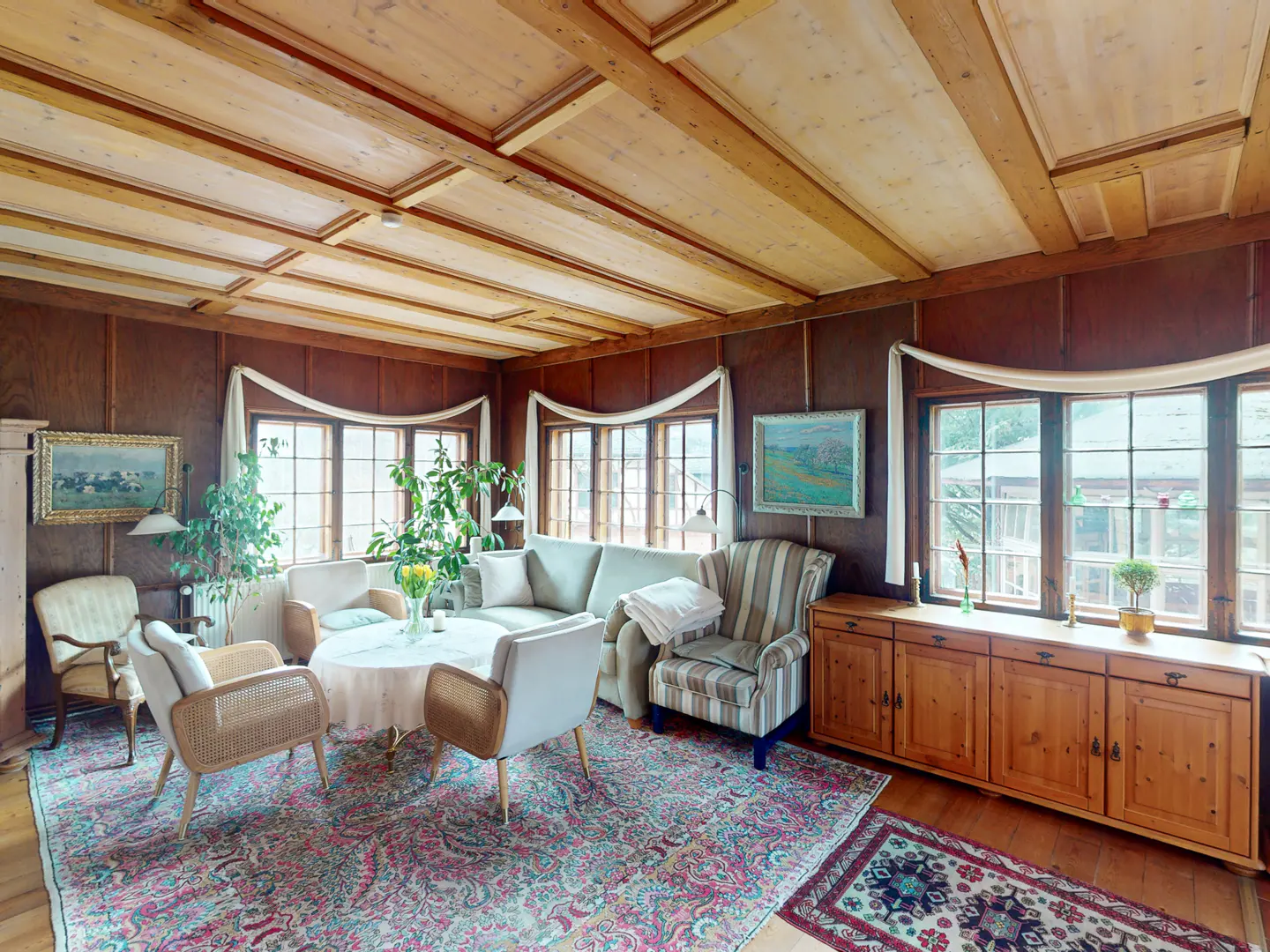 Living room with wood paneled walls, light wood ceiling, and large windows. A round table sits in the center, surrounded by chairs and a sofa. A patterned rug covers the floor.