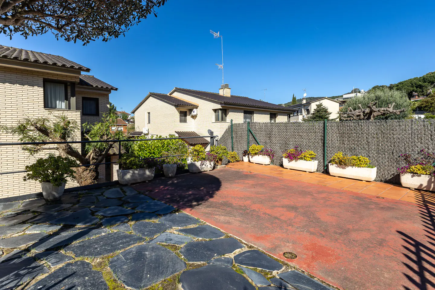 Outdoor patio with stone and red concrete flooring, potted plants, and a woven fence. Houses are visible in the background under a clear blue sky.