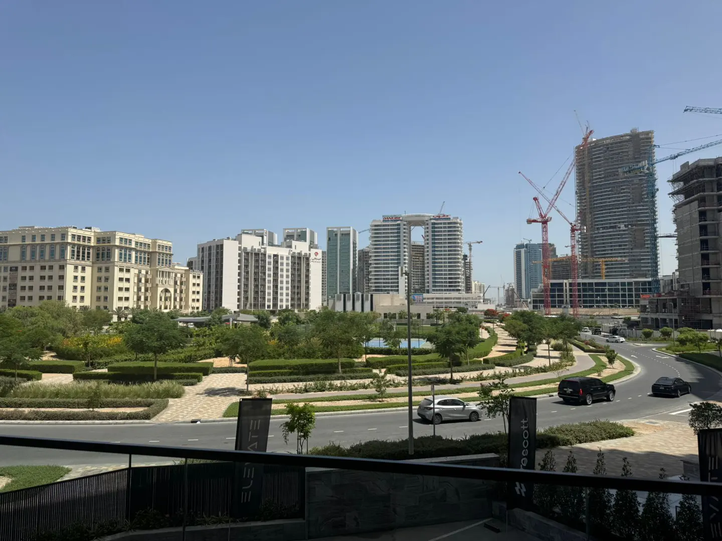 Cityscape view with modern buildings, green spaces, and roads with cars under a clear blue sky. Construction cranes are visible near taller buildings.