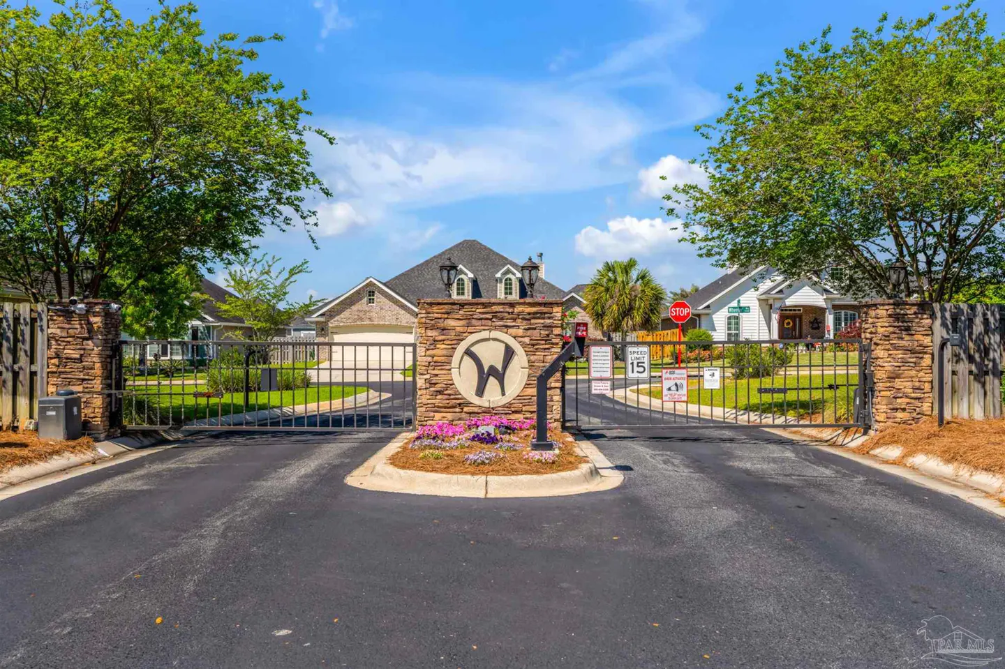 Gated community entrance with stone pillars, a 'W' logo, and flower bed. Houses visible in the background under a blue sky.