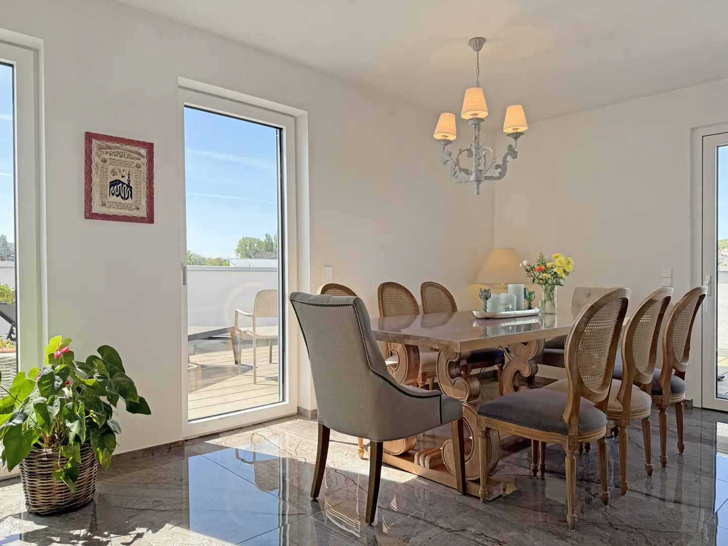 Bright dining room with a wooden table, gray chairs, and a chandelier. A plant sits near a glass door leading to a balcony.