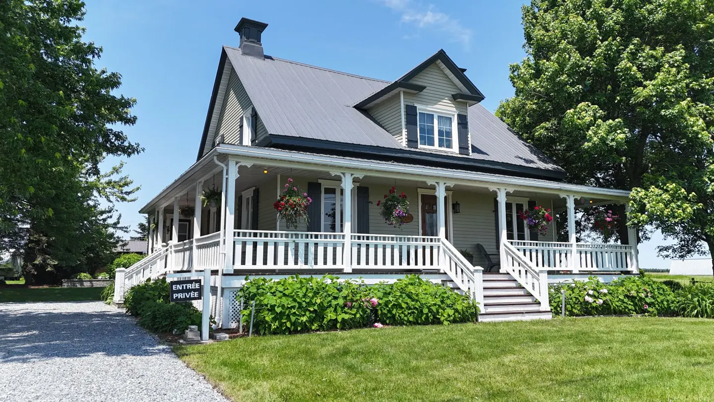 Two-story house with a gray metal roof and a white porch with hanging flower baskets. Green lawn and trees surround the house.