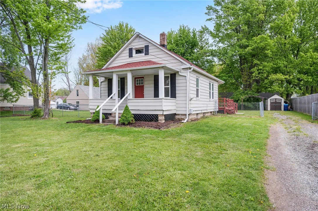 A quaint, light-gray house with a red roof and door, surrounded by a lush green lawn and trees. A gravel driveway leads to a shed in the backyard.