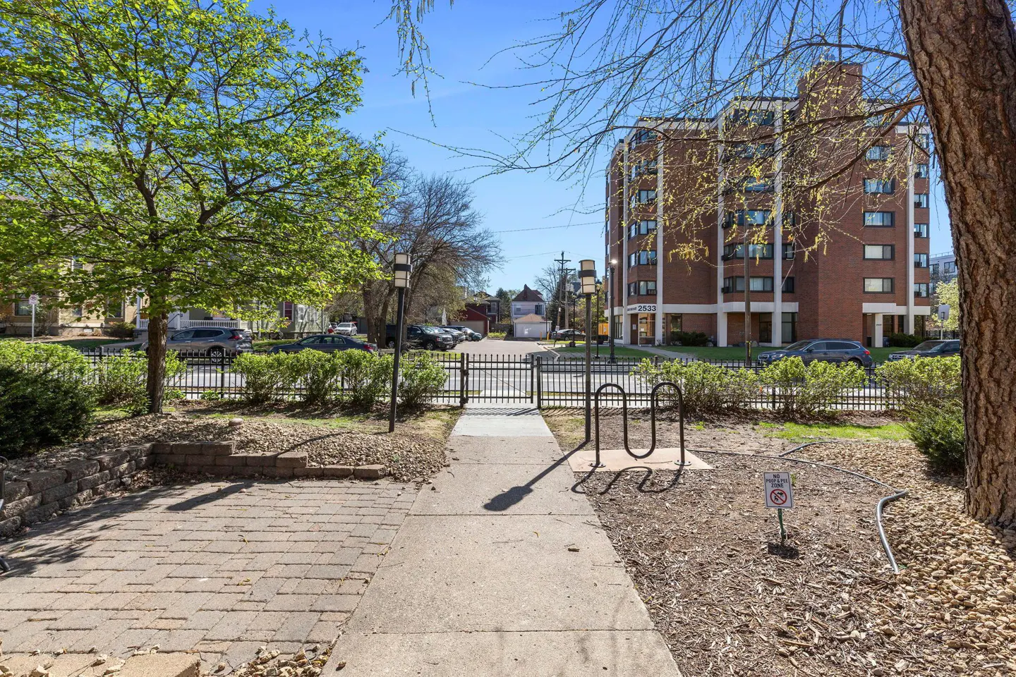 Exterior view of a brick apartment building with a sidewalk, bike rack, and trees on a sunny day.
