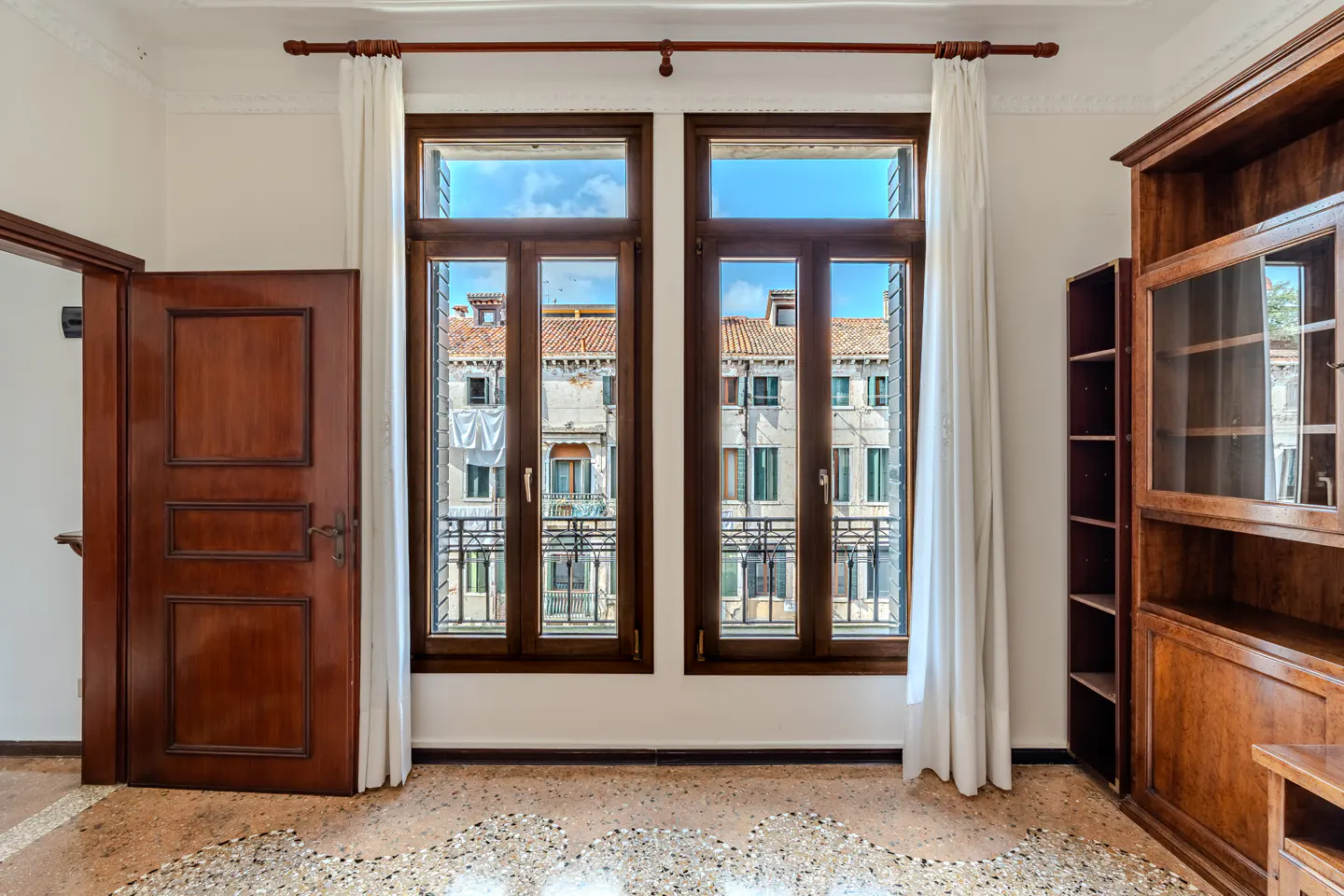 A room with two windows showing a Venice building, a wooden door, and a wooden bookcase.