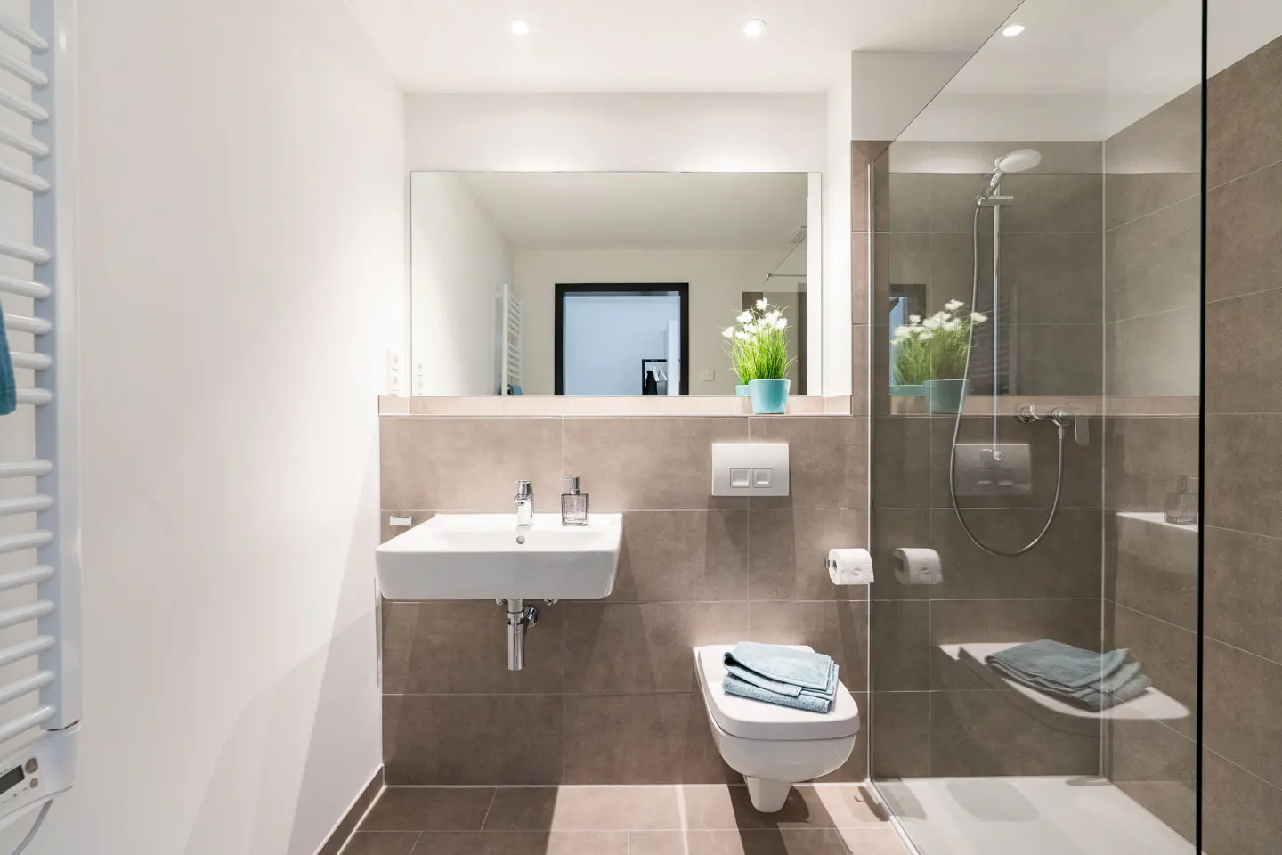 Modern bathroom with white sink, toilet with blue towels, and glass-enclosed shower with gray tile. A large mirror reflects the room.