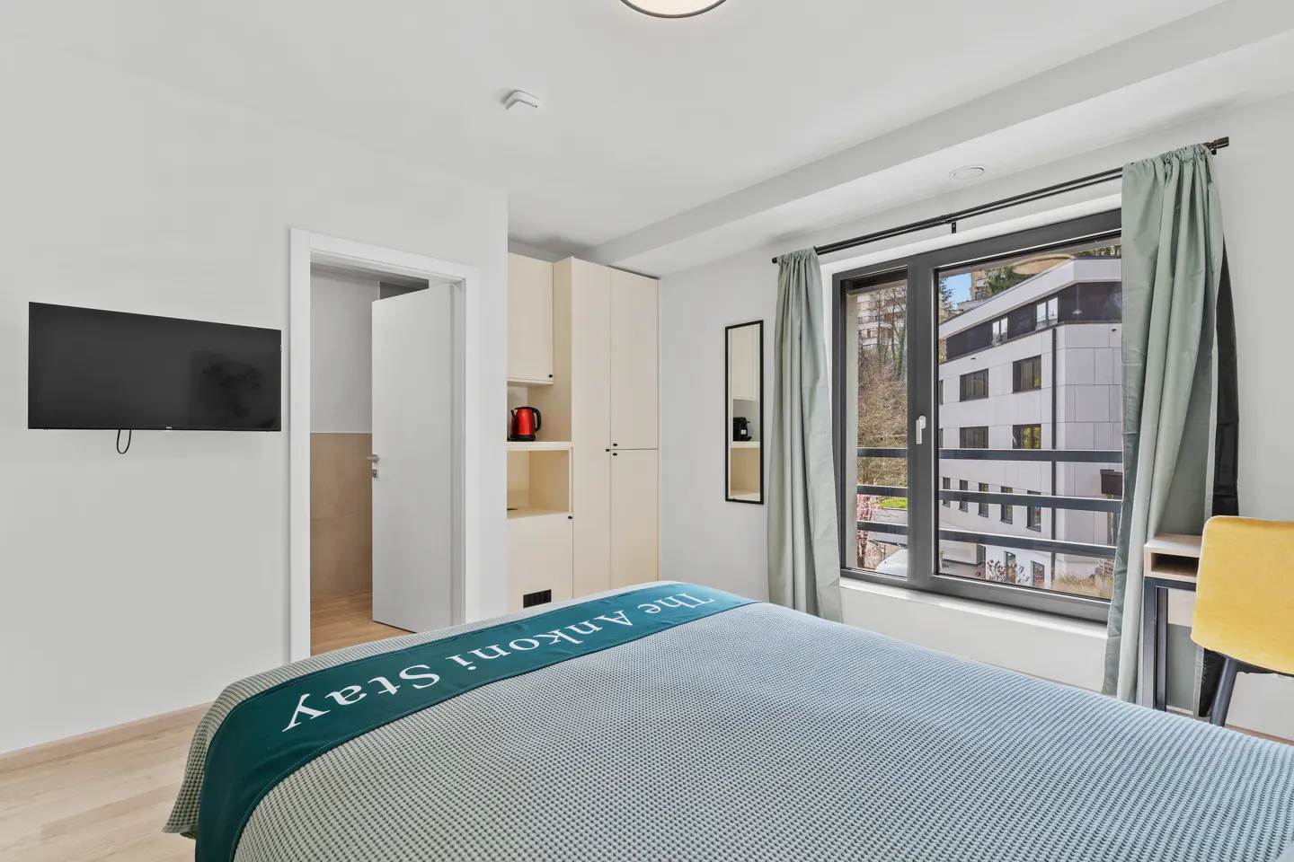 Bright hotel room with a blue bedspread, a TV, and a window with a view of a building.