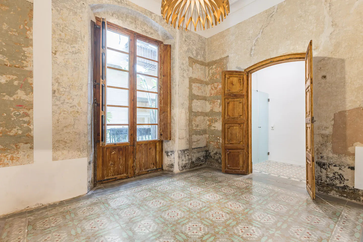 Unfurnished room with peeling walls, patterned tile floor, and wooden window and door frames. A modern wooden chandelier hangs from the ceiling.