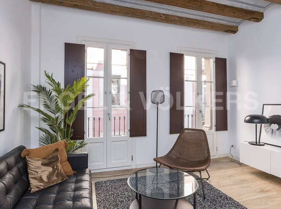 Living room with black leather sofa, brown wicker chair, glass table, and two windows with brown shutters.