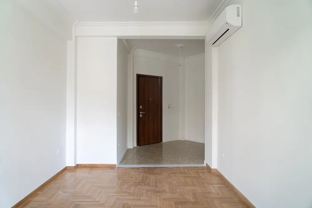 An empty room with white walls, a herringbone wood floor, and a dark wood door in the background. An air conditioner is mounted high on the wall.