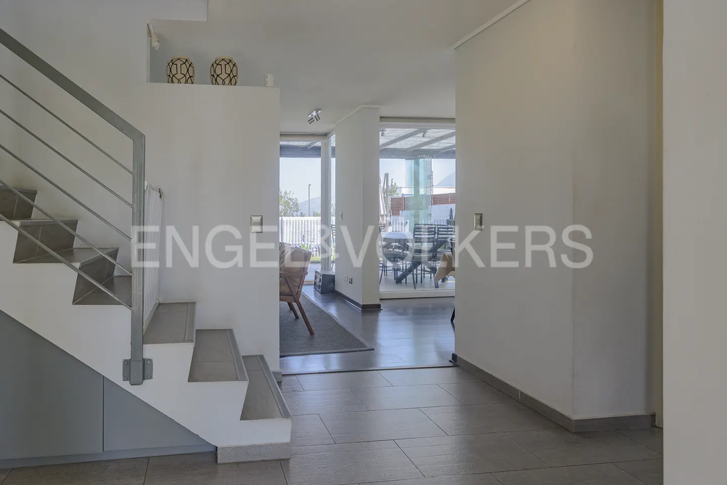 Interior view of a modern home with a staircase, tiled floor, and an open doorway to a patio with outdoor furniture.