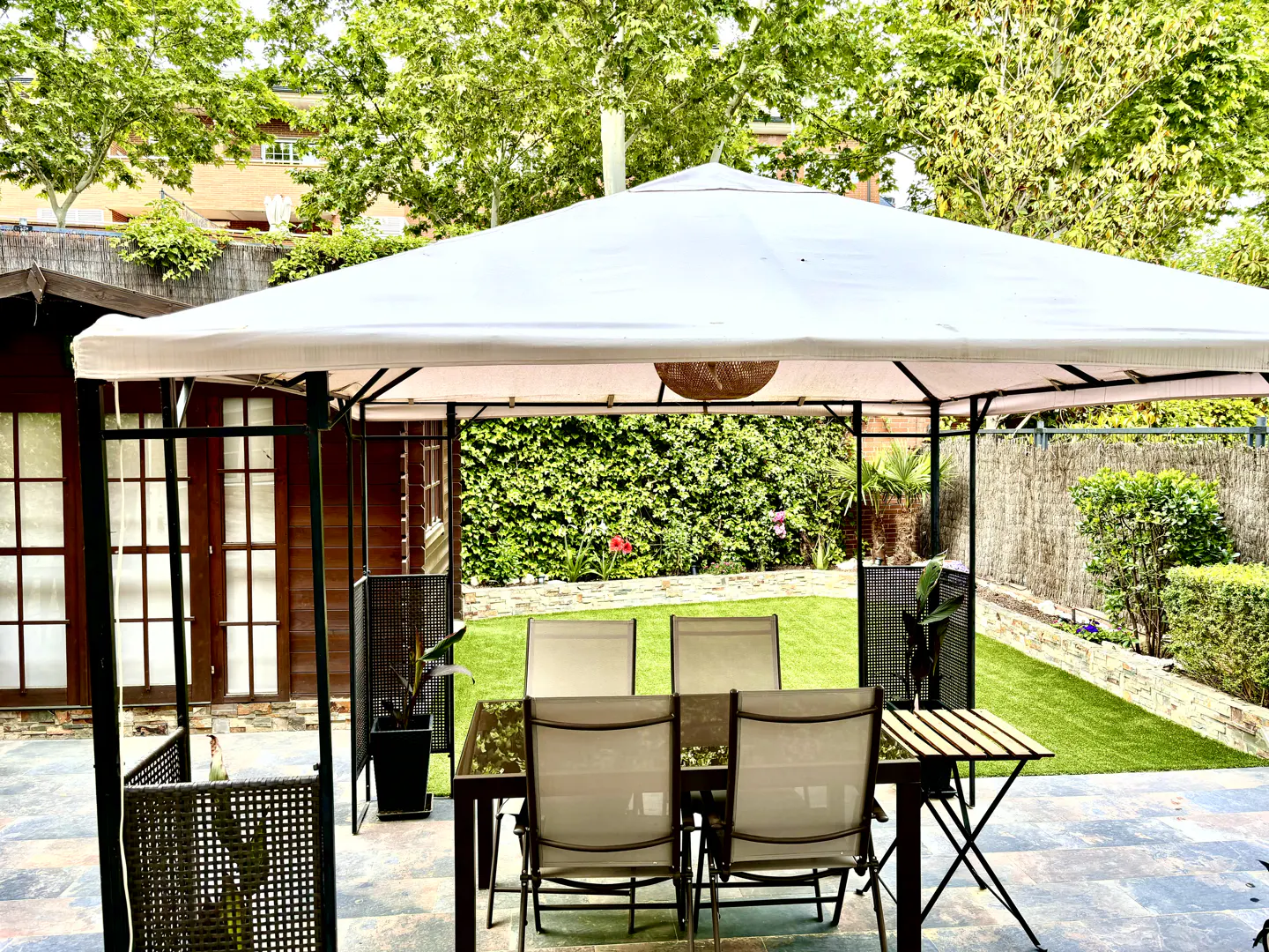 A white gazebo covers a patio dining set with four chairs on a stone floor, overlooking a lush green backyard garden.