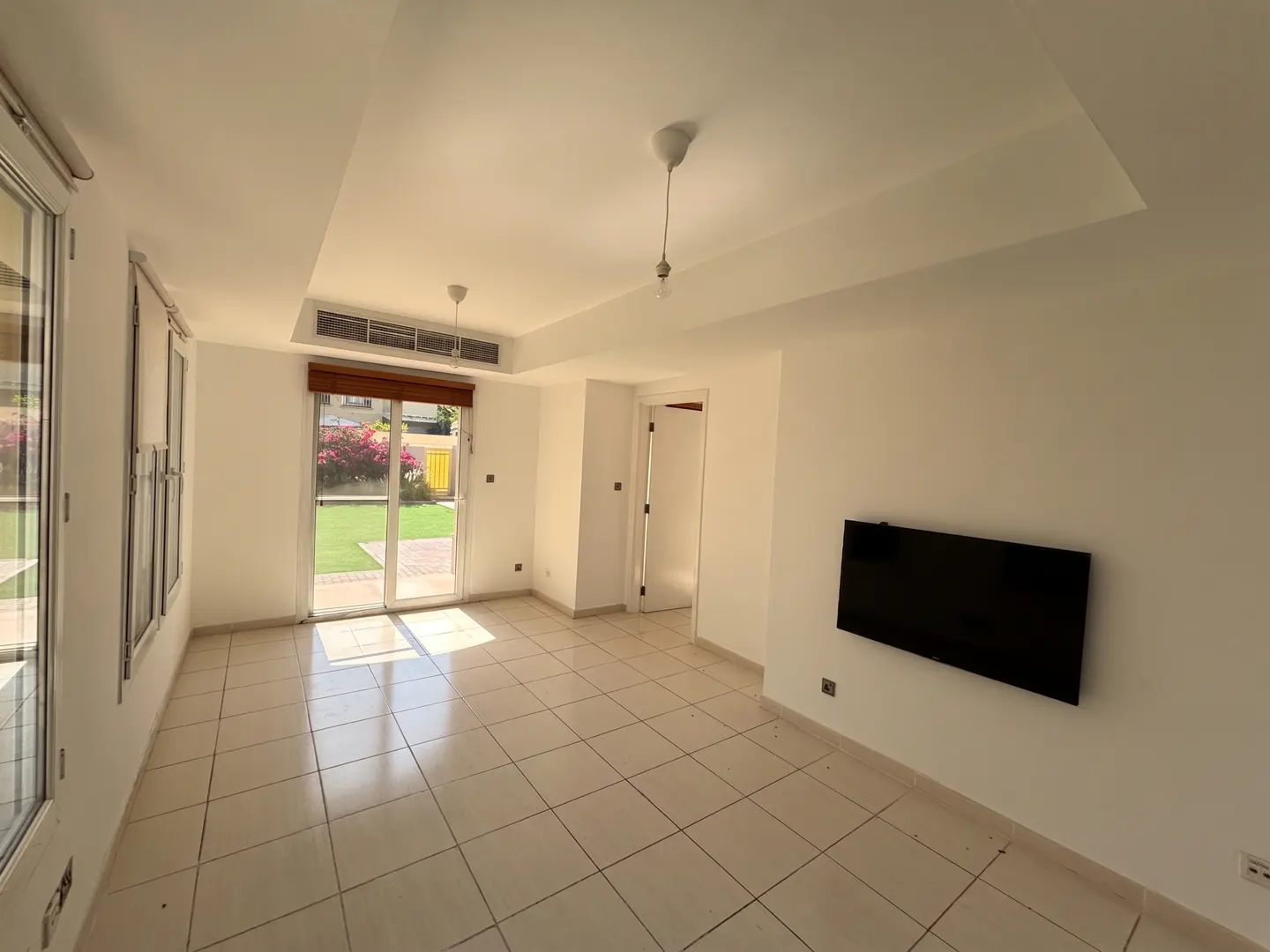 Bright, empty living room with white walls, tile floor, and a black TV mounted on the wall. Sliding glass doors lead to a green lawn.