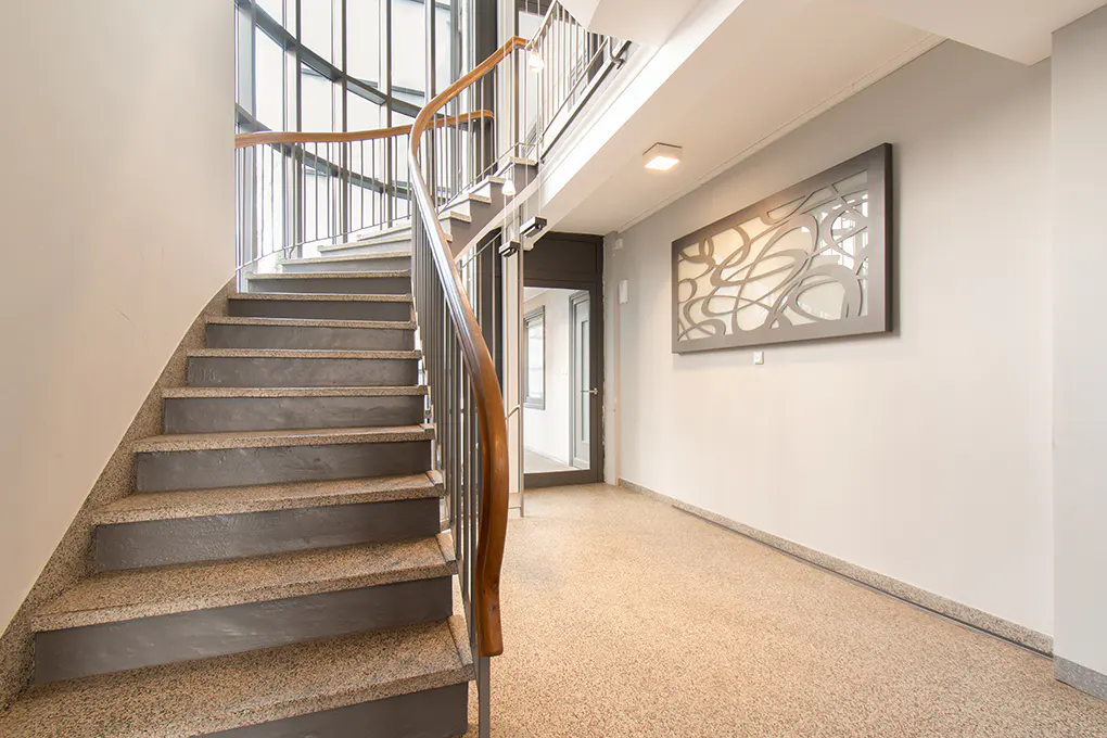 Interior view of a building with a curved staircase, wooden handrail, and a gray wall with an abstract art piece.