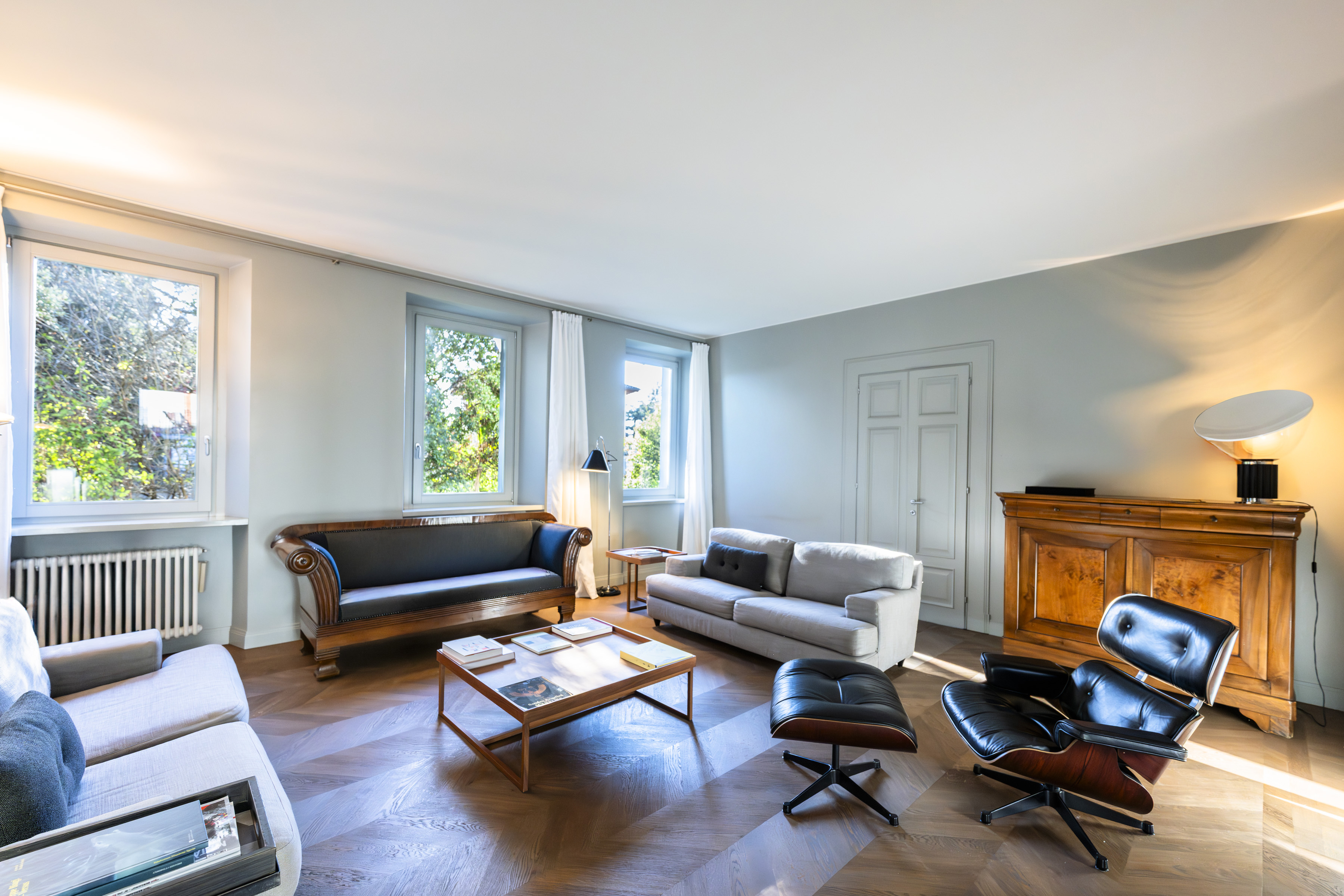 A bright living room with a black leather chair and ottoman, two sofas, and a wooden coffee table on a herringbone floor.