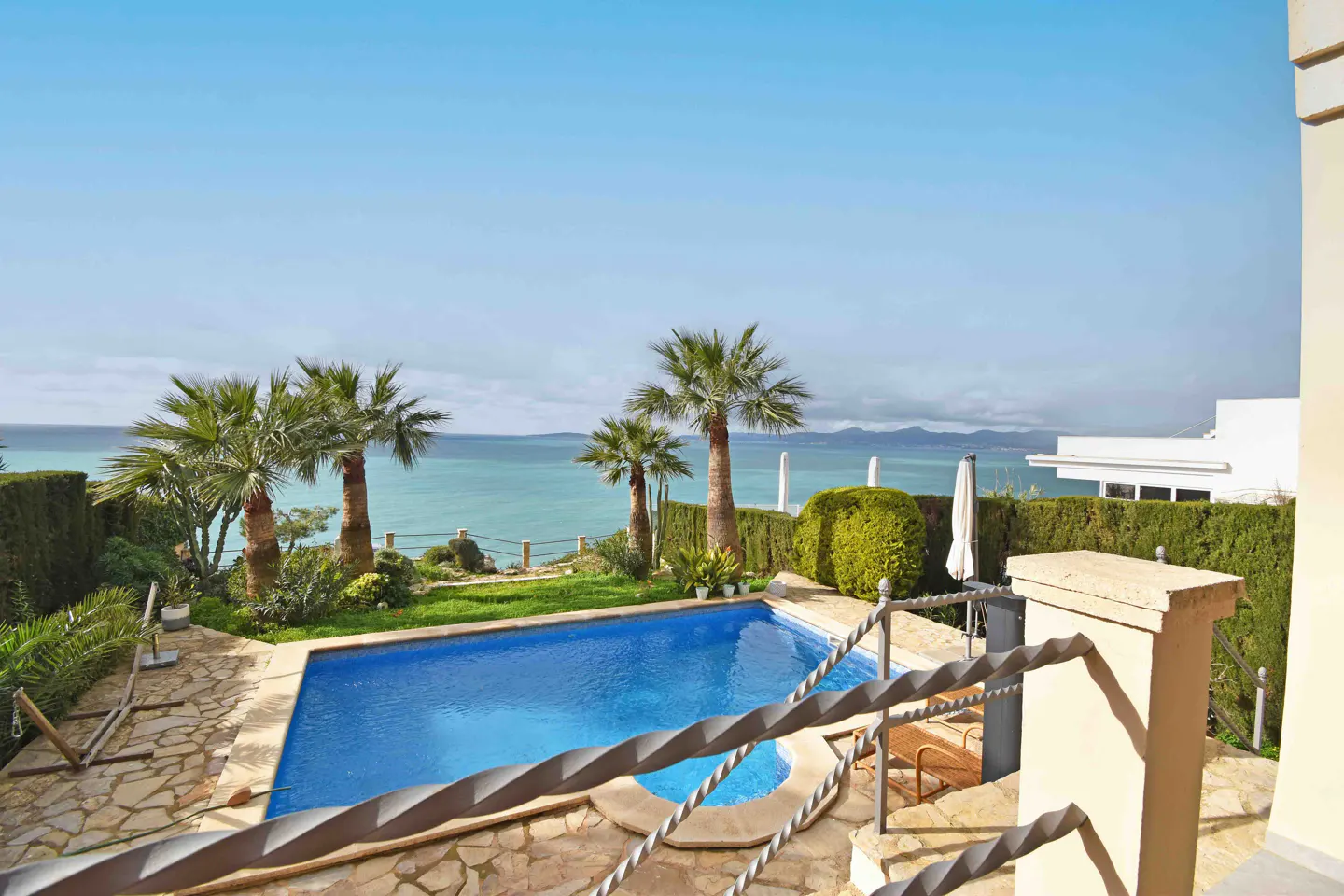 A high-angle shot shows a blue pool surrounded by palm trees and green bushes, with the ocean and sky in the background.