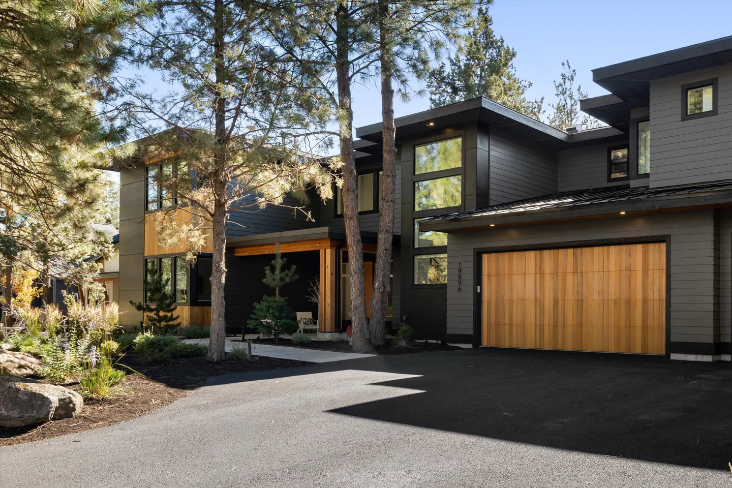 Modern two-story house with gray siding, wood accents, and a large wood garage door. Tall trees line the front of the house.
