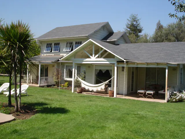 Exterior view of a two-story, light yellow house with a gray roof and a green lawn. A hammock hangs on the porch.