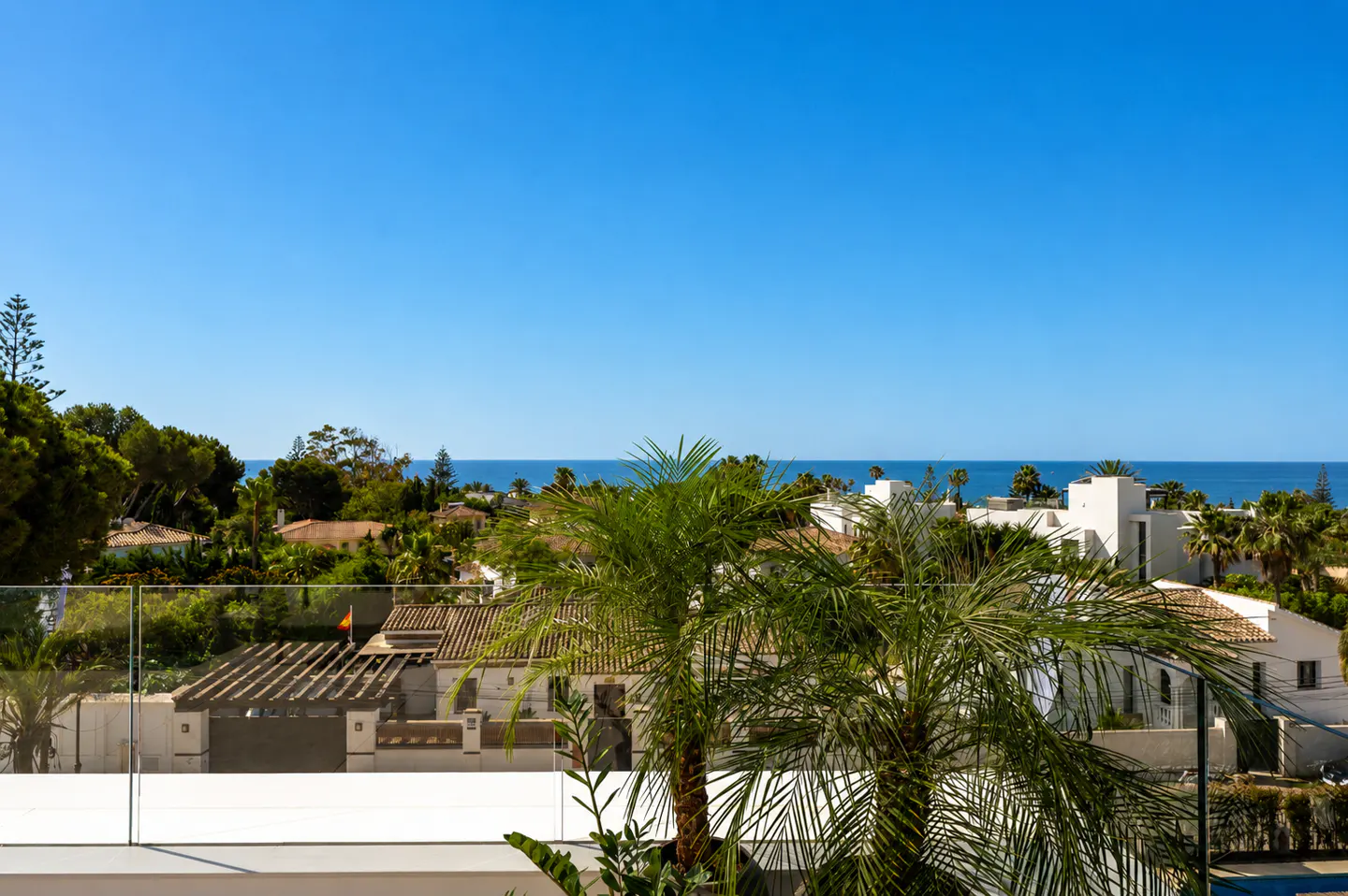 View from a balcony overlooking houses, palm trees, and the ocean under a clear blue sky.