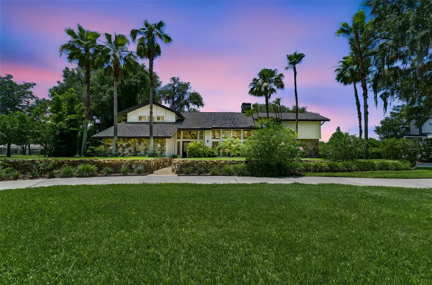 A large, two-story house with palm trees and a green lawn under a pink and blue sky.