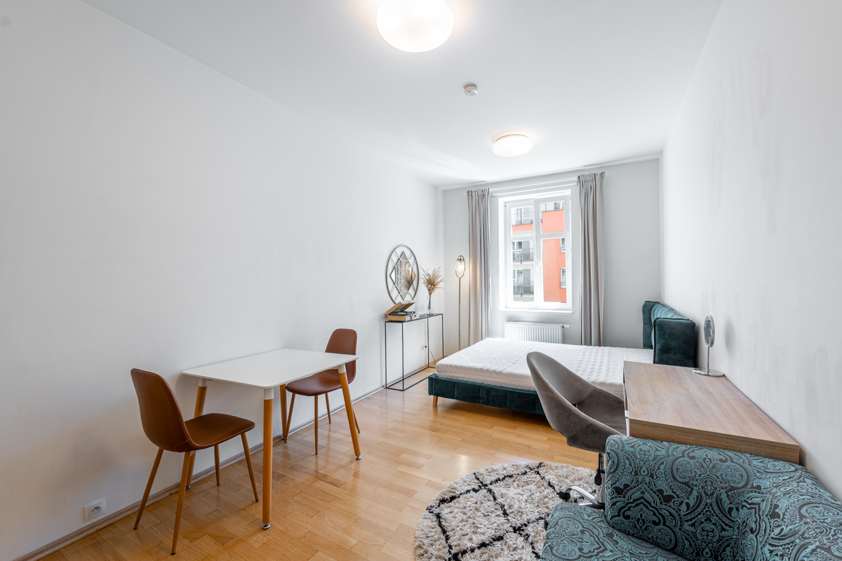 Studio apartment with a white dining table, two brown chairs, a bed with a dark headboard, and a patterned armchair. [AI-generated text]