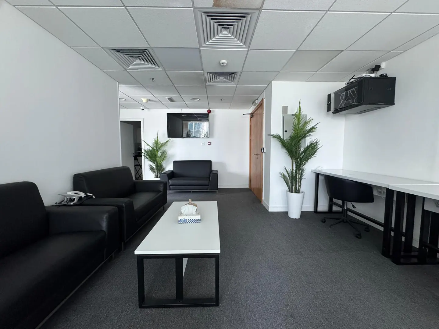 A modern office waiting area with black sofas, a white table, and a desk with a chair. Plants add a touch of nature to the space.