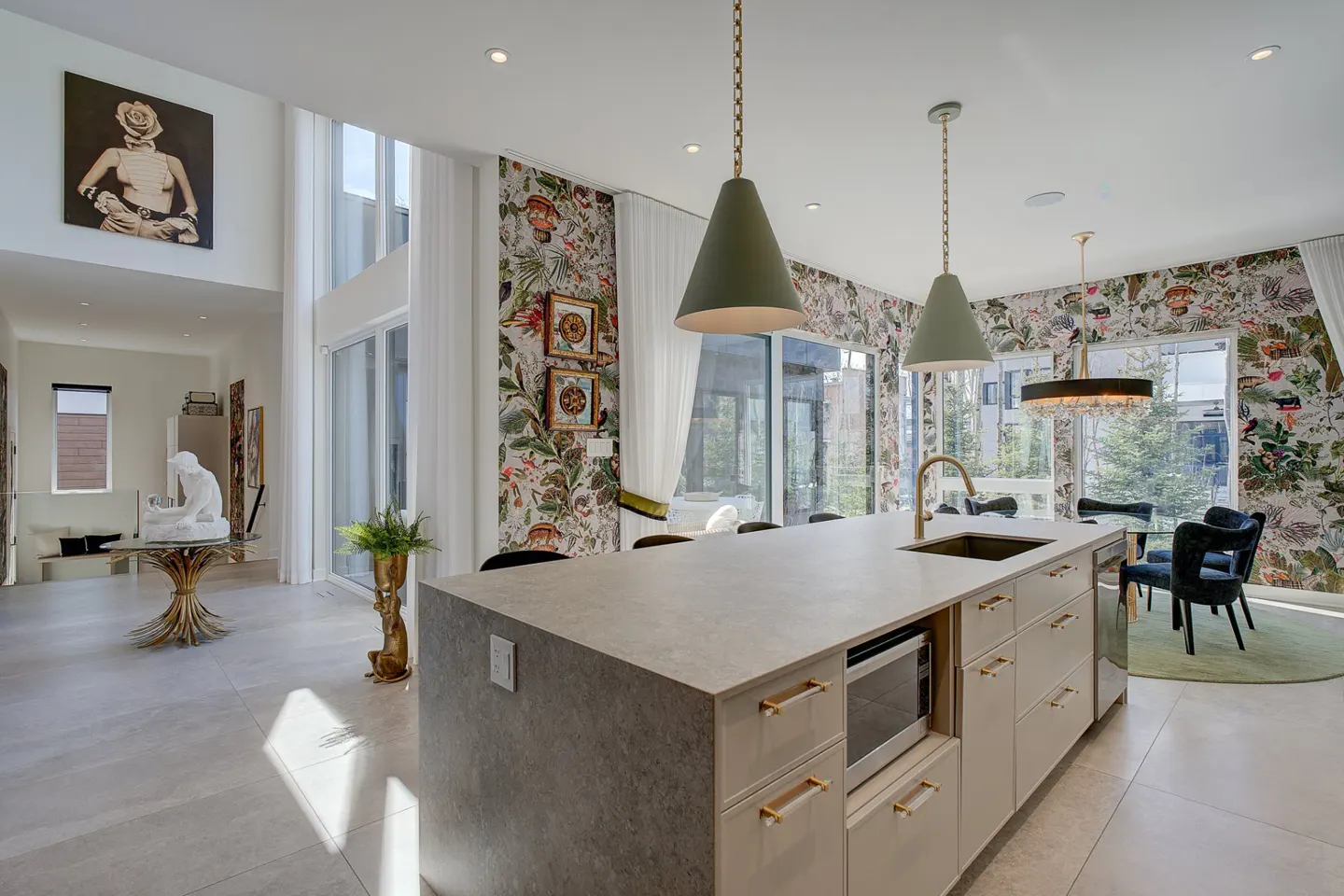 A modern kitchen with a large island, green pendant lights, and floral wallpaper. A dining area is visible in the background.
