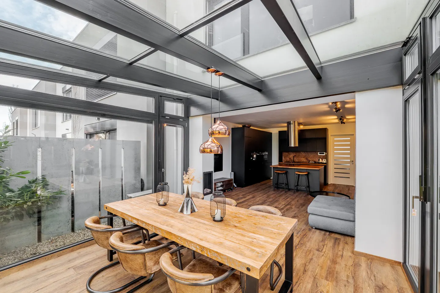 Bright dining area with a glass ceiling, a wooden table with leather chairs, and a view into a modern black kitchen.