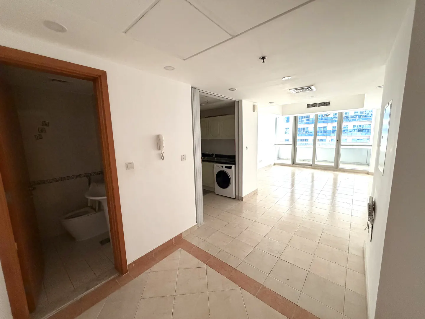 A bright, empty apartment with white walls, tiled floors, and a view of city buildings through large windows. A bathroom and kitchen are visible.