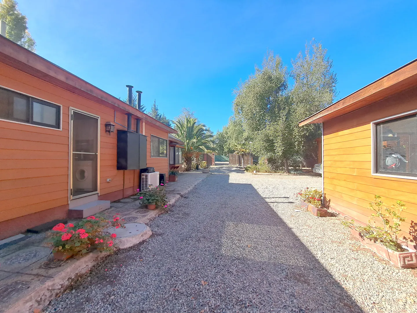 Exterior view of two orange wooden buildings with a gravel driveway and green trees under a blue sky.