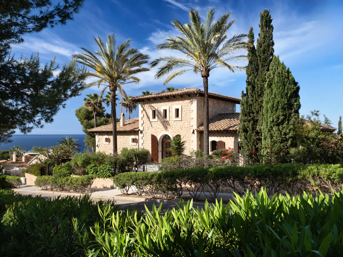 Stone house with a red tile roof, palm trees, and green foliage against a blue sky.