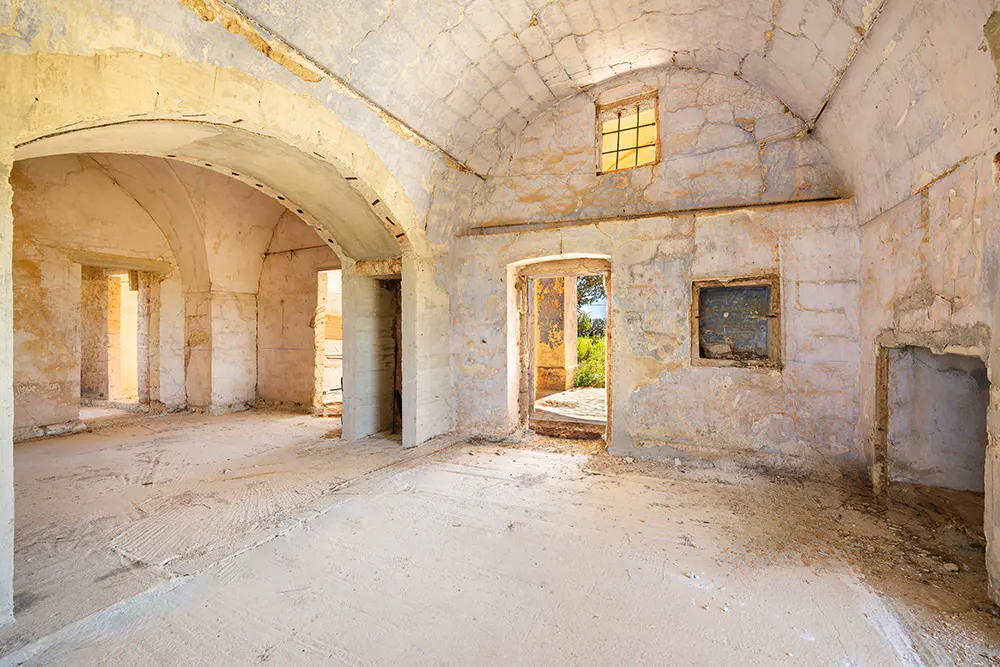 Interior view of a stone building under renovation, featuring arched doorways, small windows, and unfinished walls.
