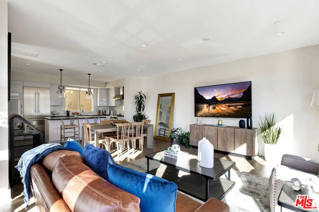 Bright, open-concept living space with a brown leather couch, blue pillows, and a TV displaying a mountain scene. A kitchen and dining area are visible in the background.