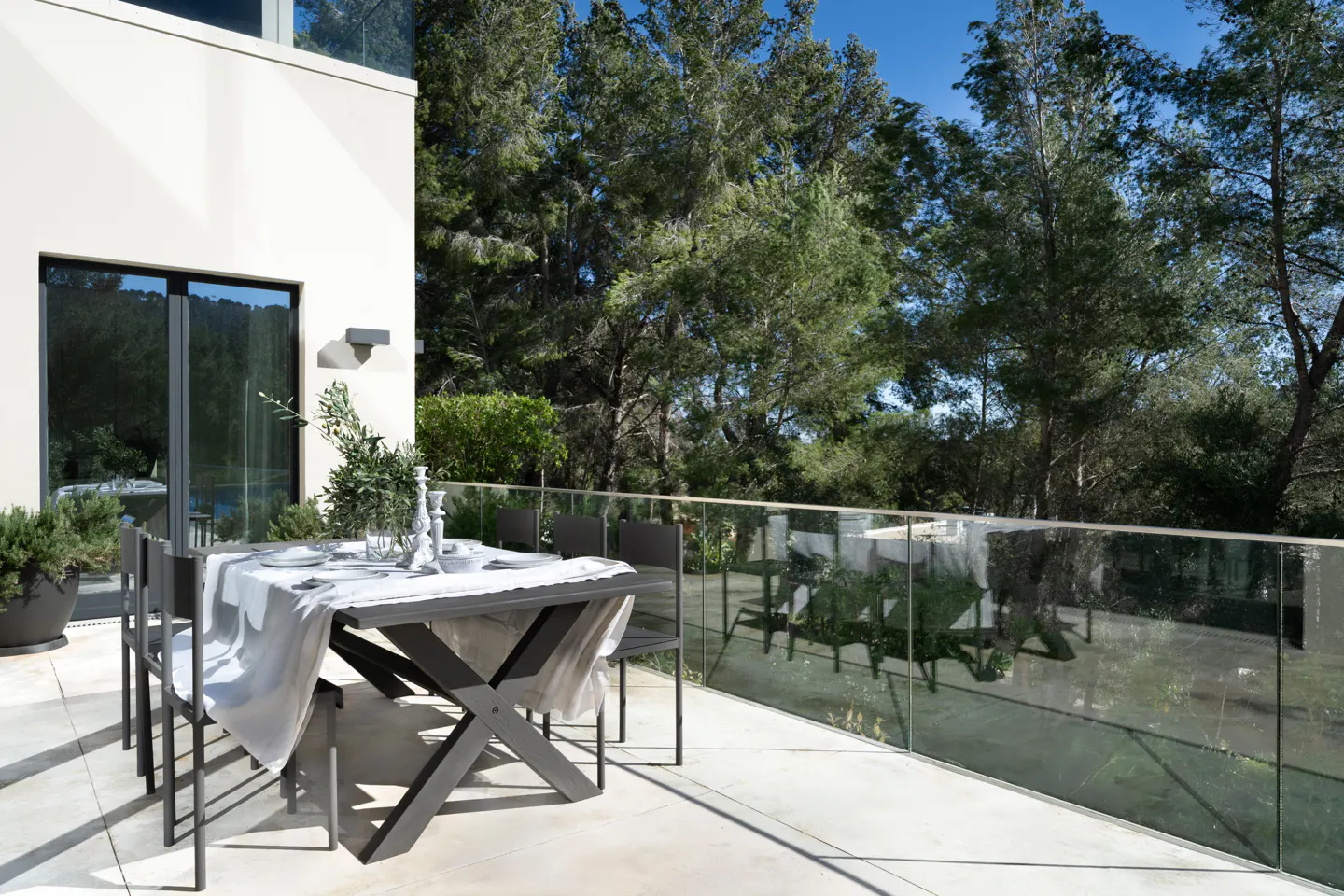 Outdoor patio with a gray table set for dining, featuring a white linen cloth, plates, and candlesticks. A glass railing overlooks lush green trees.